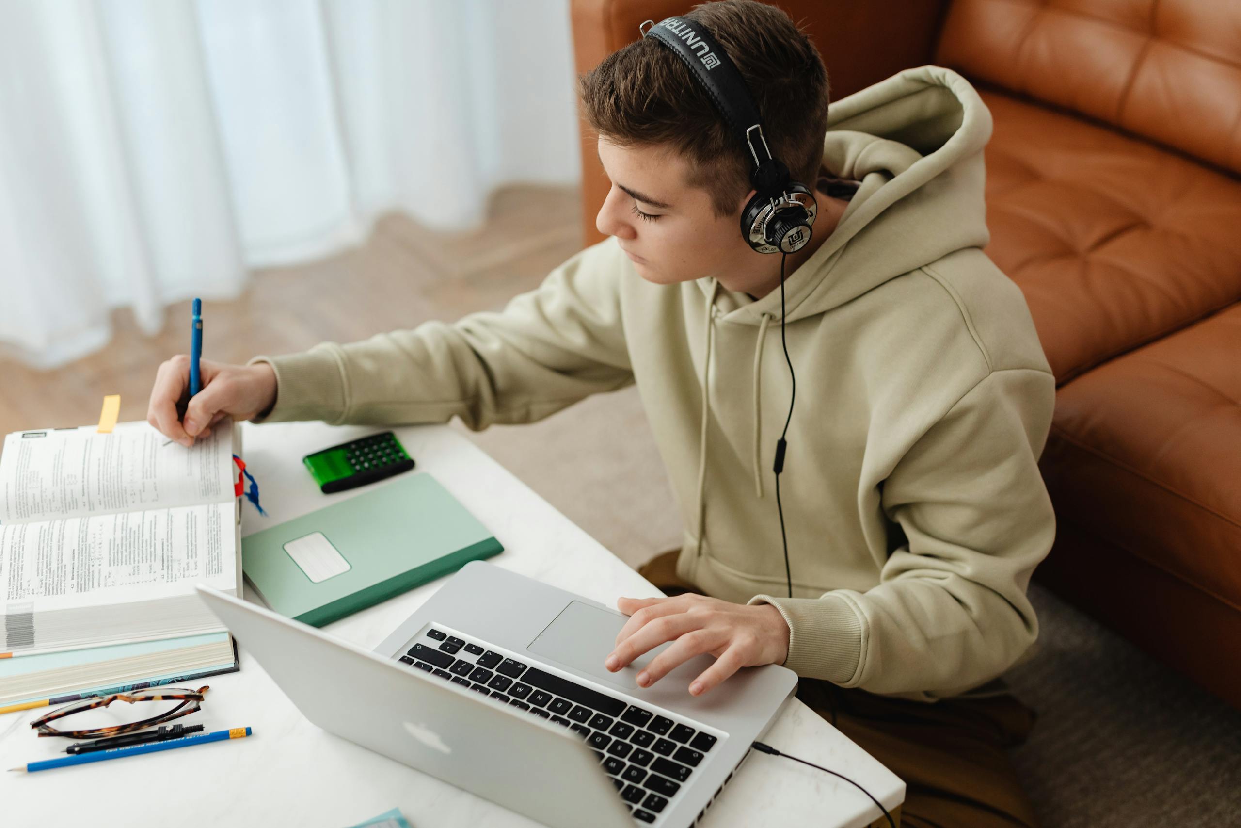 Teen studying at home using laptop and books, wearing headphones, sitting in a cozy room.