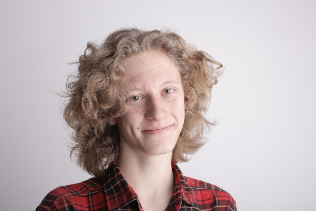 Optimistic smiling teenage guy in casual red checkered shirt with blond long curly hair looking at camera while standing against gray background