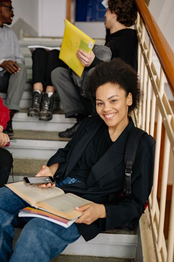 Smiling student sitting on stairs, holding a book and phone, showcasing a relaxed study moment.