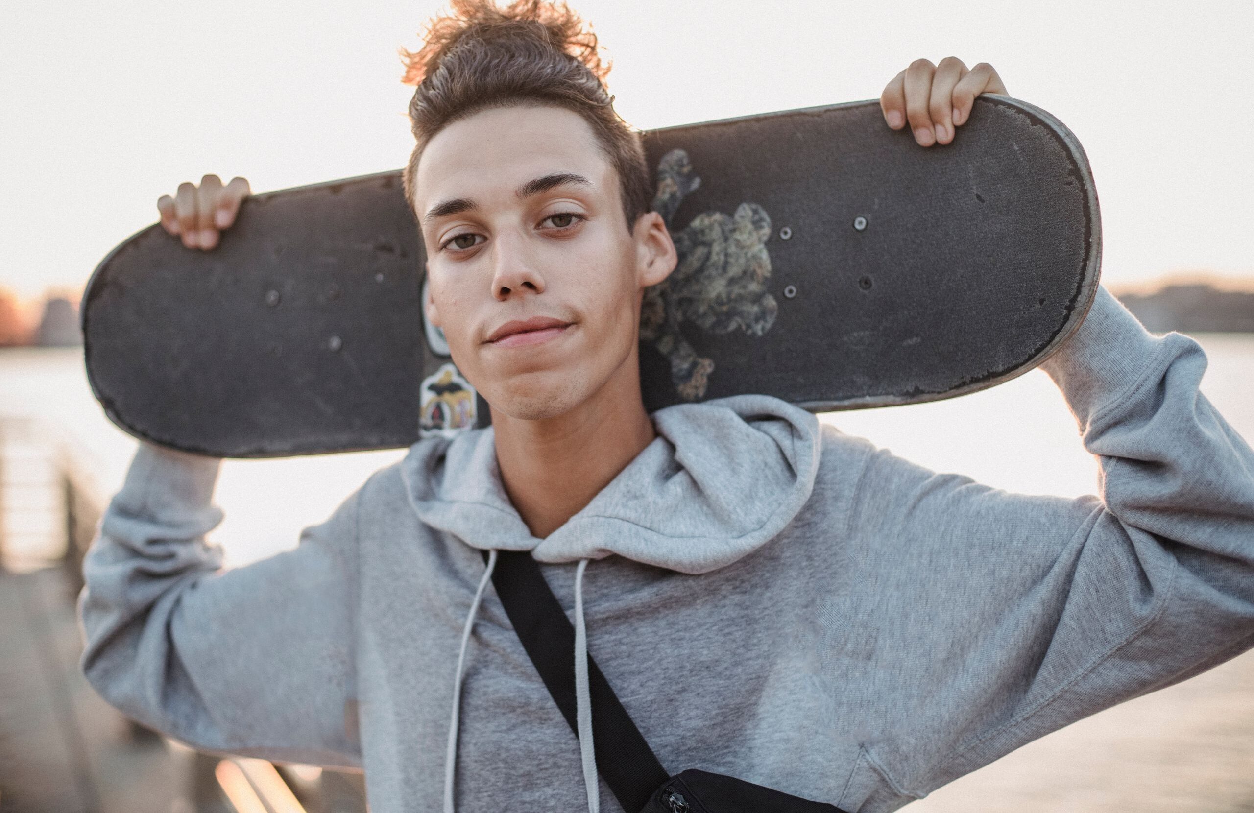 Serious young man in hoodie carrying skateboard on shoulders and looking at camera
