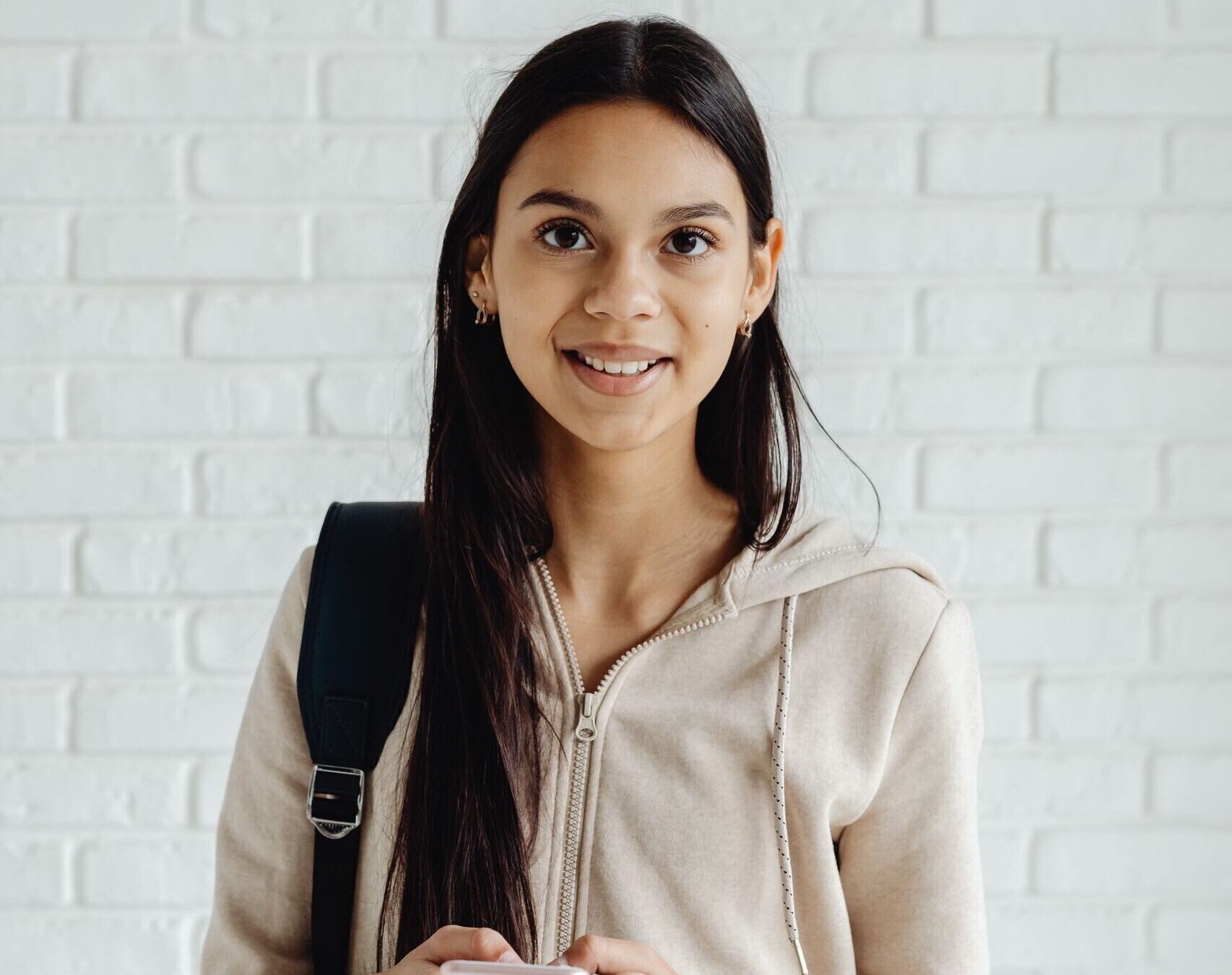 Smiling teenager in hoodie with smartphone and backpack against white brick wall.
